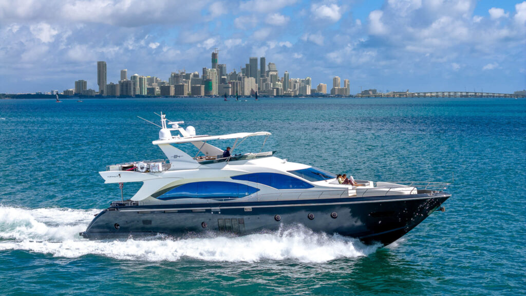 Miami Rent A Yacht A large white and blue yacht moves through blue water with a city skyline and bridge visible in the background under a partly cloudy sky. Miami Yacht Rental